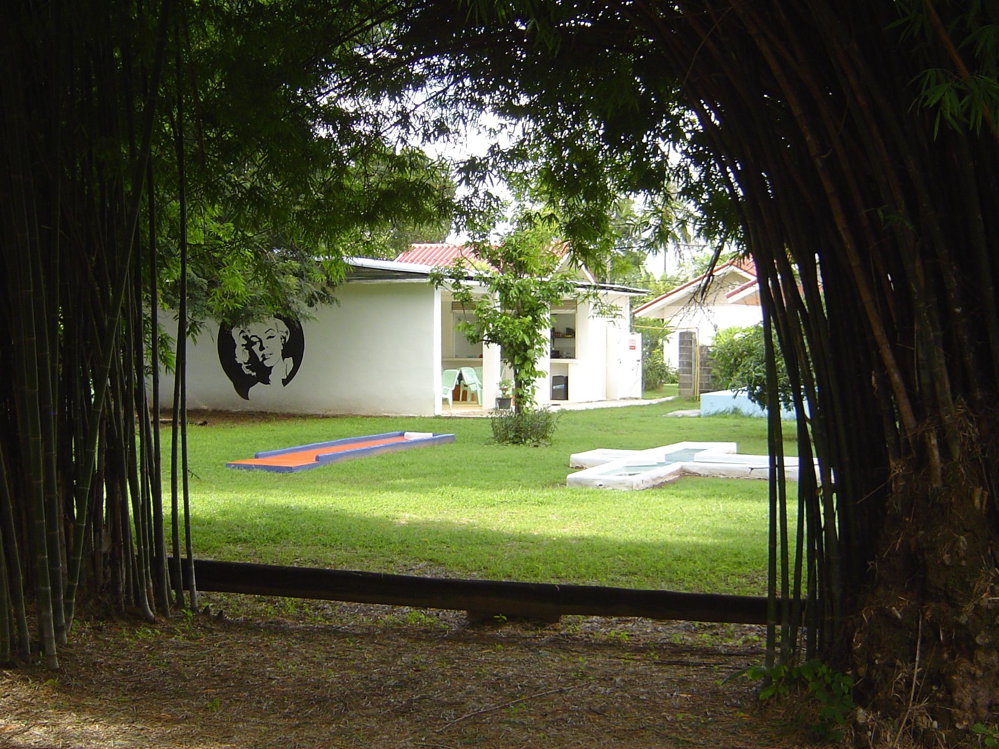 A view through a bamboo archway of a courtyard with a mural, a small pool, and a building with a red roof.