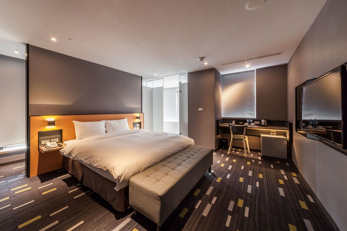 A modern hotel room with a large bed, bedside table, and a desk with a chair.