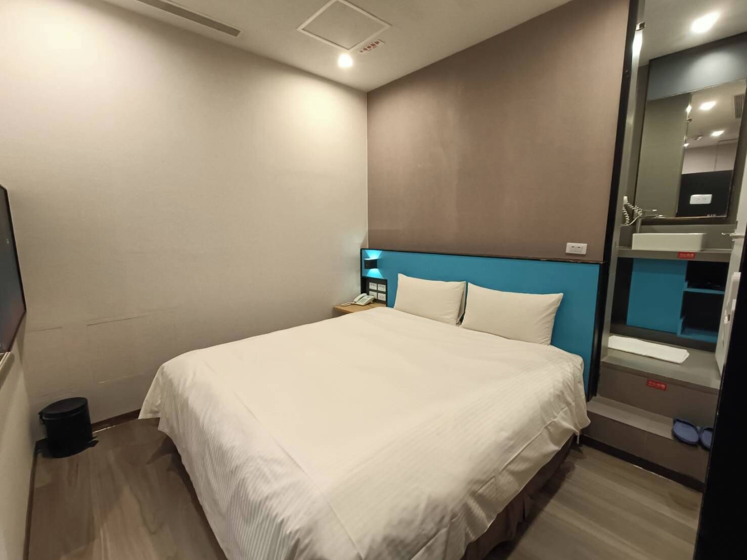 A modern hotel room with a large bed, a nightstand, a mirror, and a built-in shelf with toiletries.