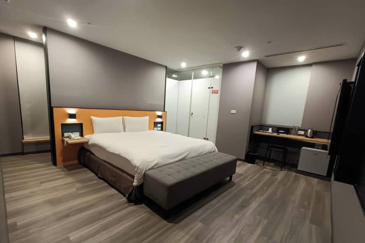 Quarto de hotel moderno com uma cama grande, mesas de cabeceira, uma escrivaninha e uma kitchenette pequena.