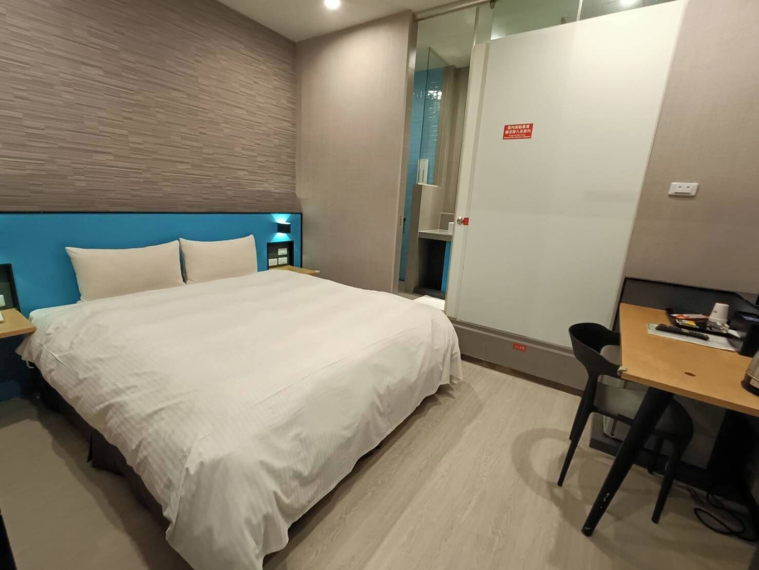 Habitació d'hotel amb llit, escriptori i cadira. Una paret blava i un capçal de llit de fusta.