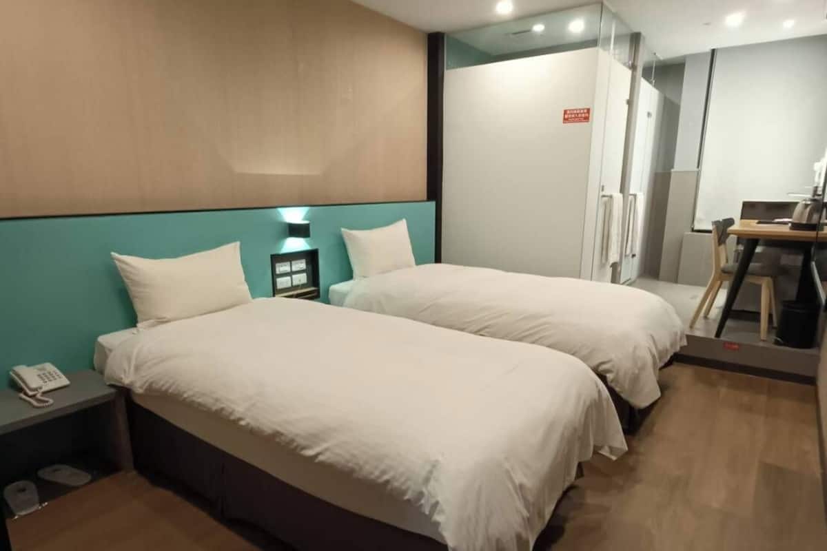 Quarto de hotel com duas camas, uma escrivaninha, uma cadeira, um telefone e uma pequena lixeira.