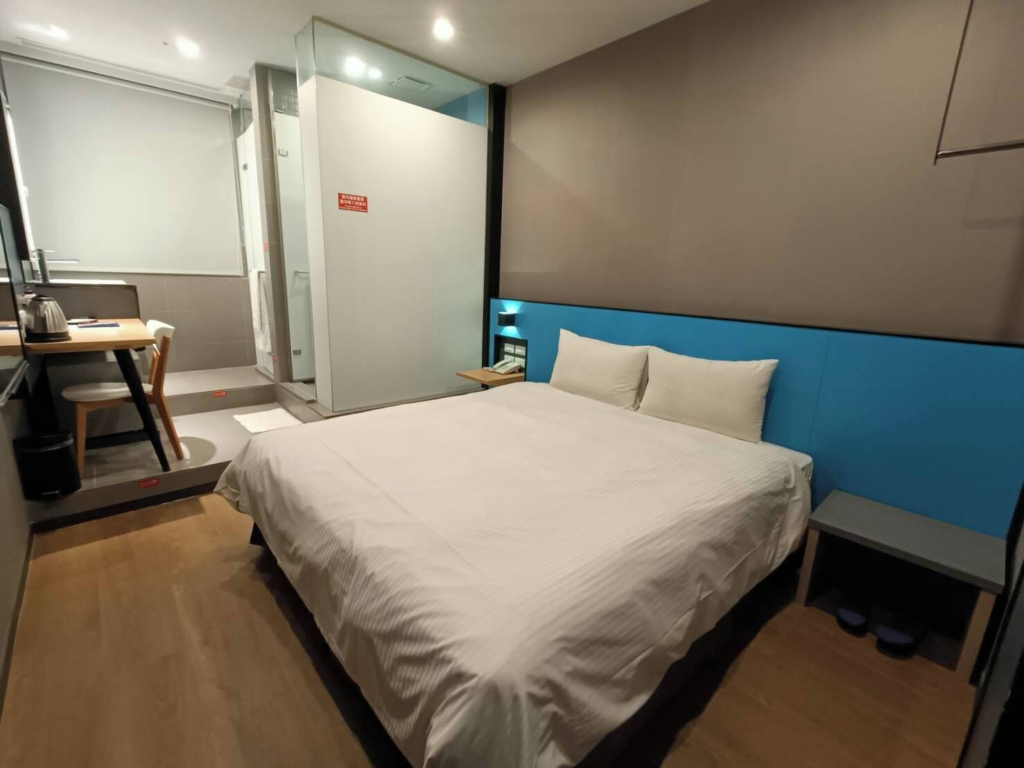 A modern hotel room with a large bed, a desk with a chair, a small table, and a nightstand with a lamp.