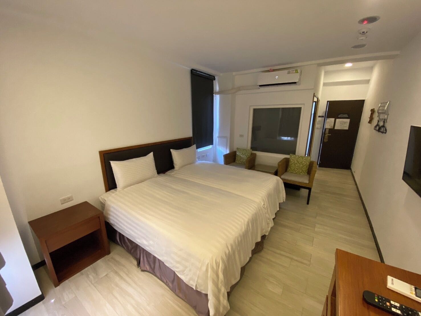 A hotel room with a large bed, a bedside table, a chair, a TV, and an air conditioning unit.