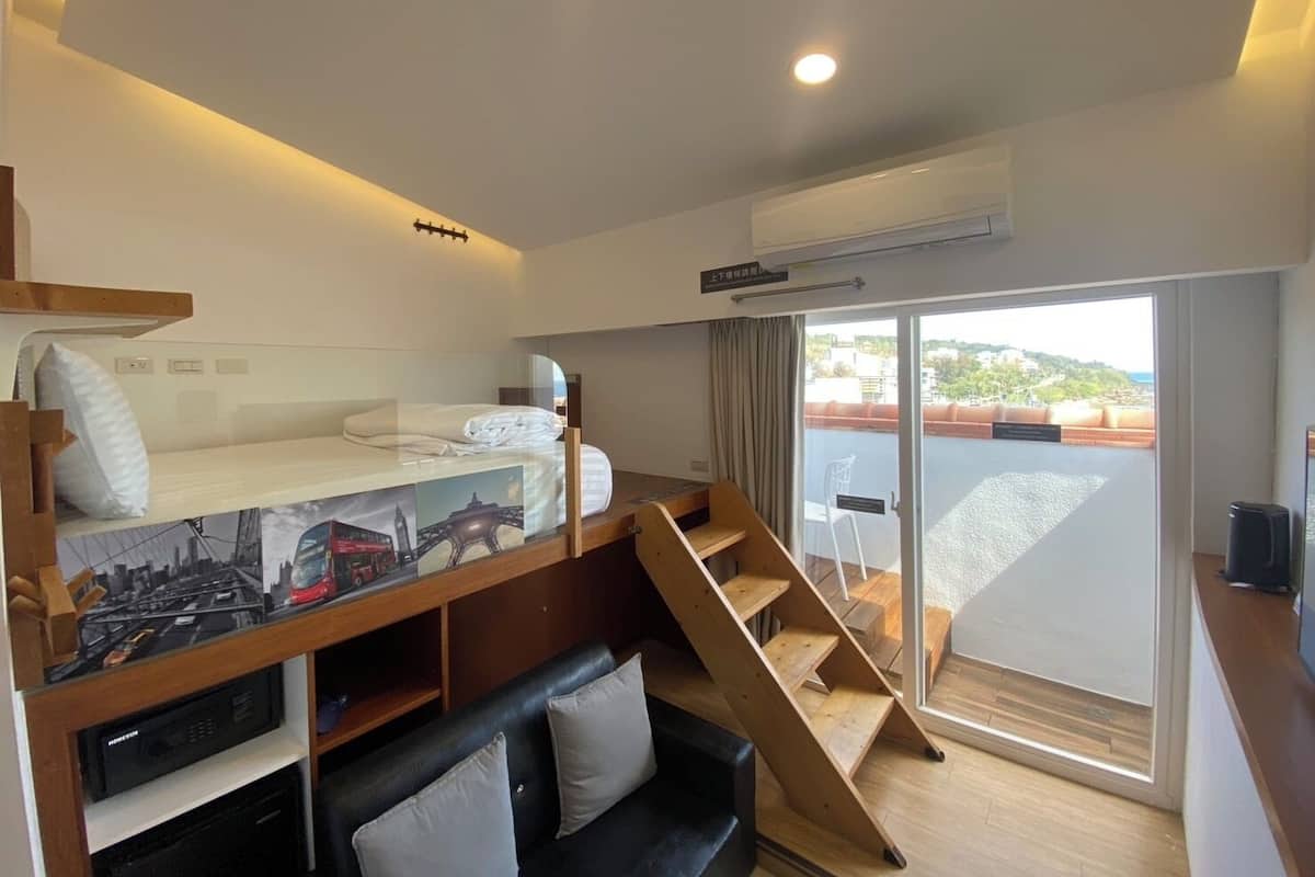 A compact hotel room with a bunk bed, a small sofa, a staircase, and a sliding glass door leading to an outdoor balcony.