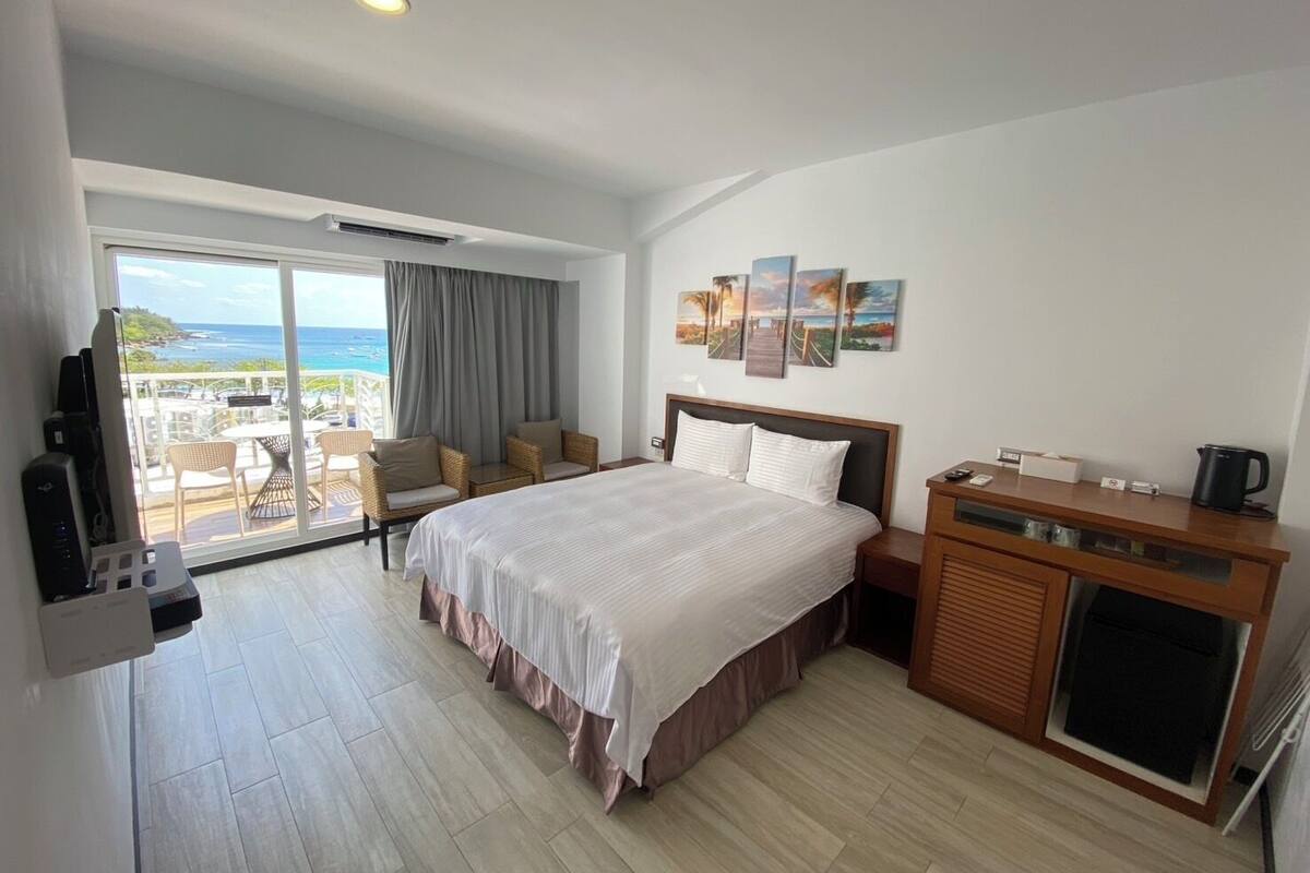 A hotel room with a large bed, a desk, a chair, a TV, and a view of the ocean.
