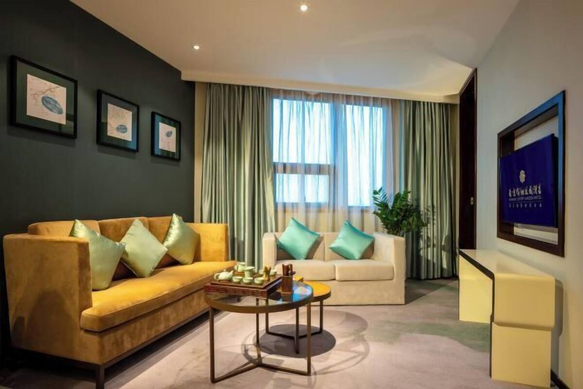Une chambre d’hôtel moderne avec un canapé jaune, un canapé blanc et une petite table ronde sur laquelle se trouve un plateau de collations. Des rideaux sont accrochés aux fenêtres, un tableau encadré est accroché au mur et un panneau bleu se trouve à droite.
