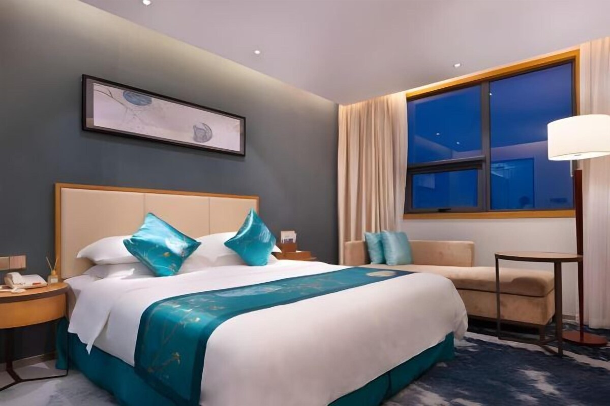 Habitación de hotel con una cama grande, una mesita de noche con lámpara, una pequeña mesa y una ventana con cortinas.