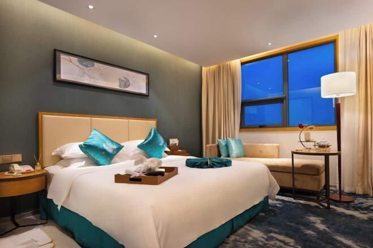 Une chambre d’hôtel avec un grand lit, une table de chevet avec une lampe, une petite table avec un vase et une fenêtre avec des rideaux.
