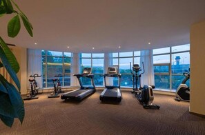 Een sportschool met loopbanden, hometrainers en een elliptische machine, grote ramen en een plant.