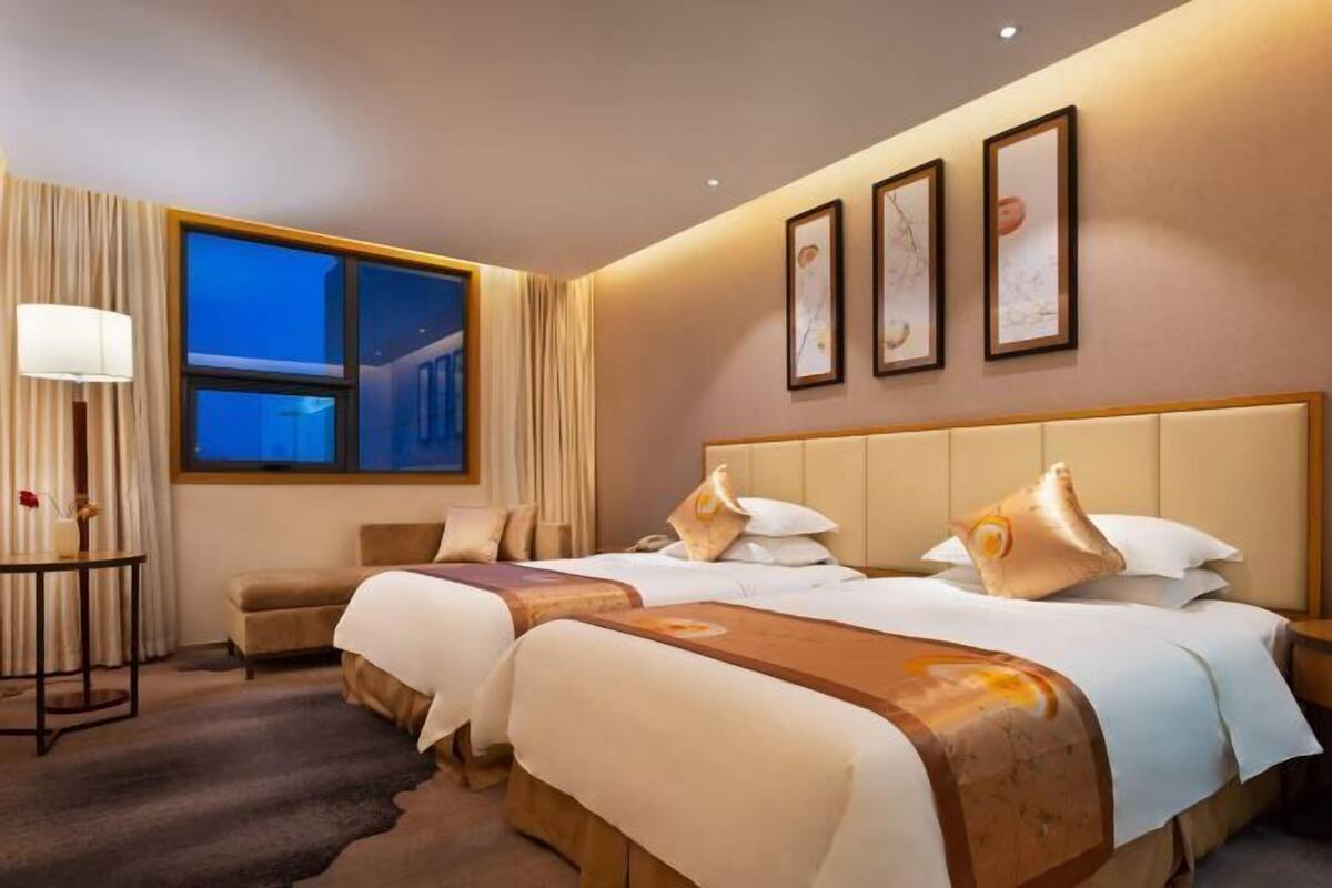 Une chambre d’hôtel avec deux lits, une table de chevet, une lampe, une fenêtre avec des rideaux et des œuvres d’art encadrées accrochées au mur.