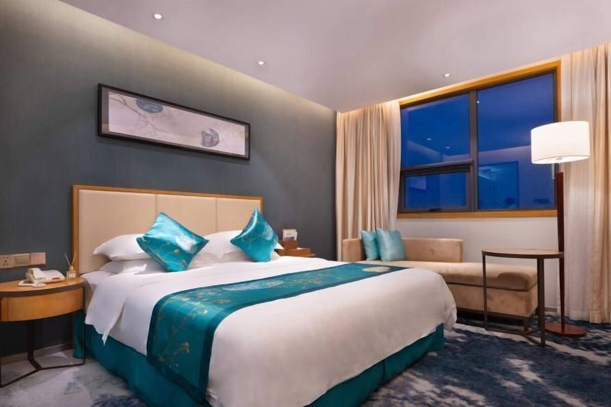 Une chambre d’hôtel avec un grand lit, un bureau, une chaise, une table de chevet, une lampe et une fenêtre avec des rideaux.