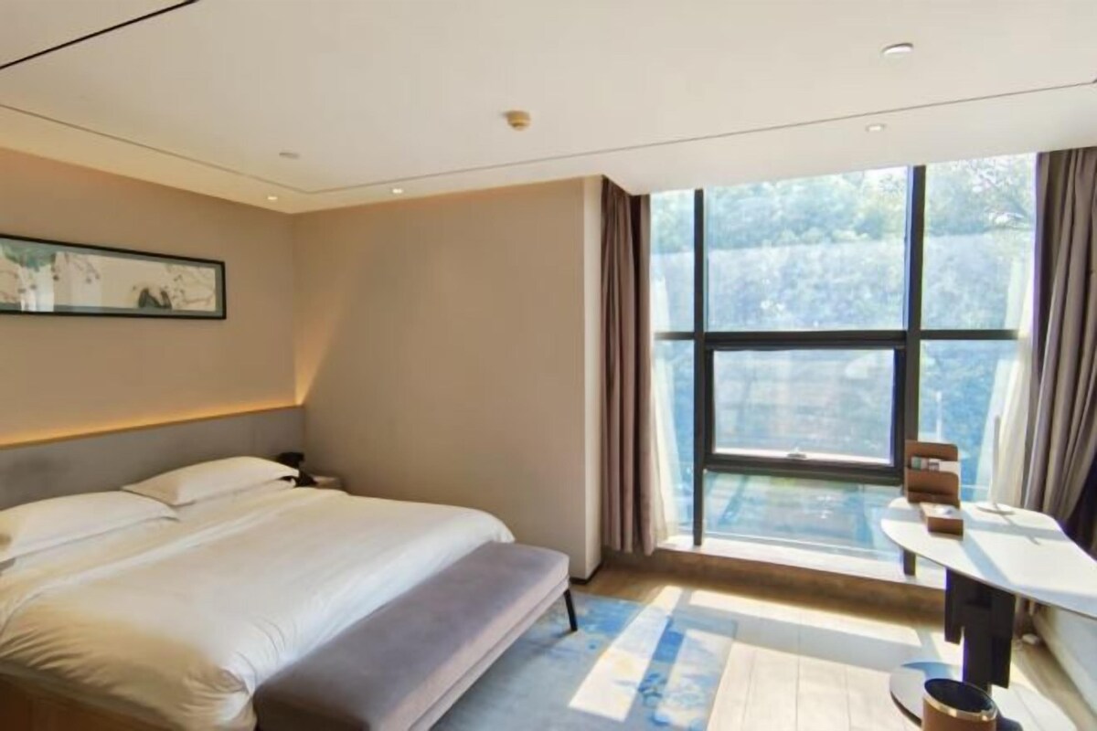 Una habitación de hotel moderna con un ventanal grande, una cama, una mesita de noche y un escritorio pequeño.