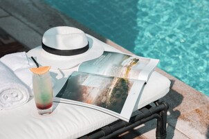 Un área de descanso junto a la piscina con una silla lounge blanca, un sombrero blanco, un vaso de bebida helada y una revista.