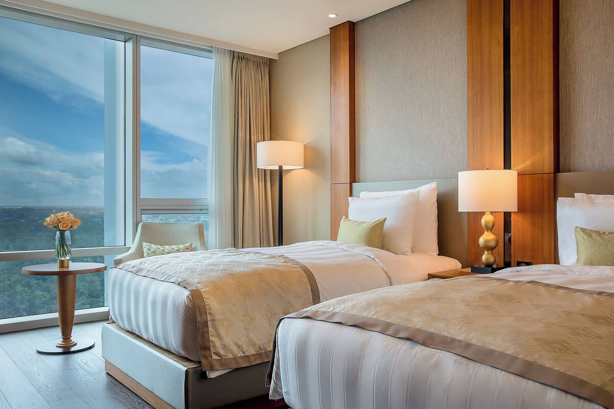 A hotel room with two beds, a large window, a vase with flowers, and a view of the city.