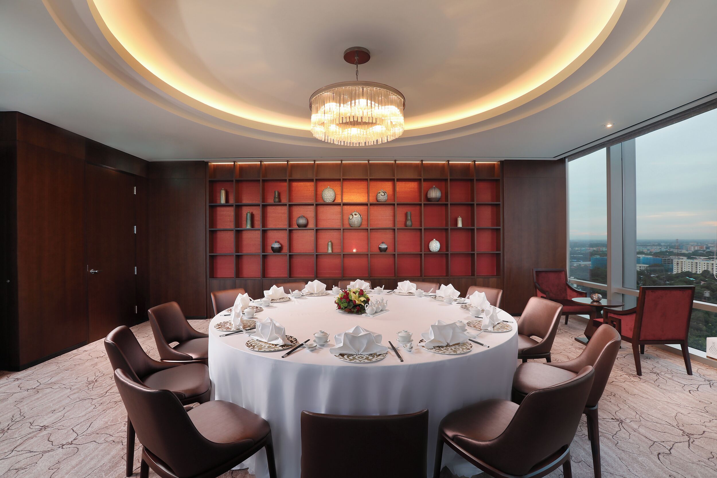 A round dining table with white tablecloth, surrounded by brown chairs, set for a meal. A chandelier hangs from the ceiling, and a wall of decorative vases is visible.