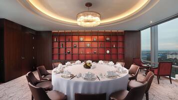 A round dining table with white tablecloth, surrounded by brown chairs, set for a meal. A chandelier hangs from the ceiling, and a wall of decorative vases is visible.