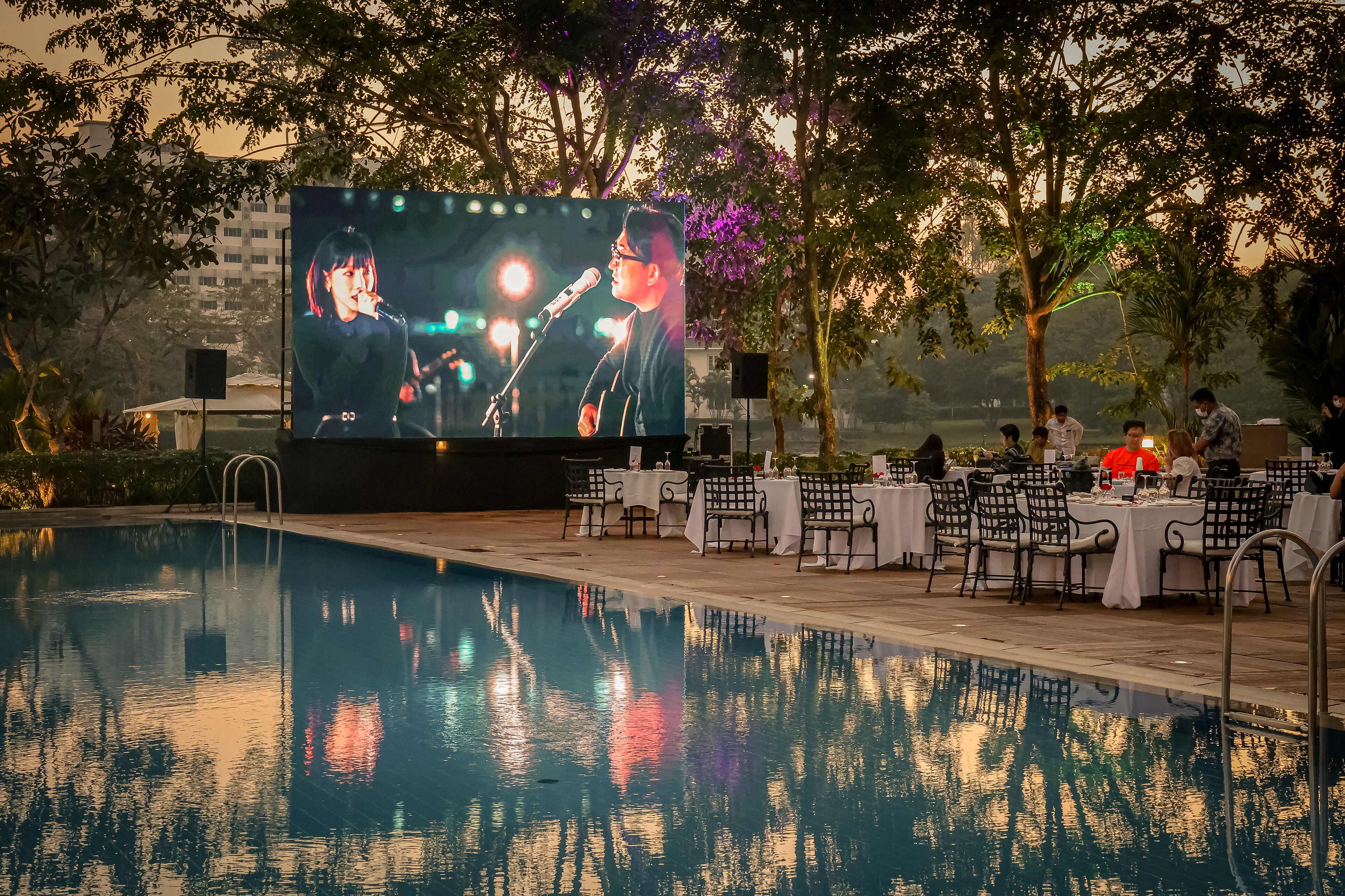 A poolside area with a large screen displaying a performer, surrounded by tables and chairs.