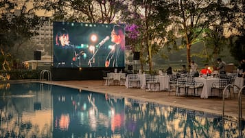 A poolside area with a large screen displaying a performer, surrounded by tables and chairs.