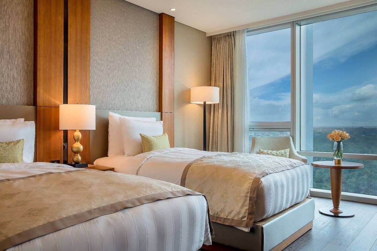 A hotel room with two beds, a large window with a city view, and a small table with a vase of flowers.