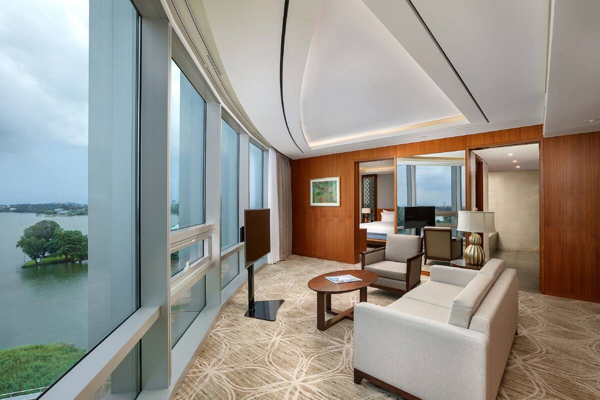A modern hotel room with a large window offering a view of a lake and trees, a cozy seating area with a sofa and armchairs, and a wooden accent wall.