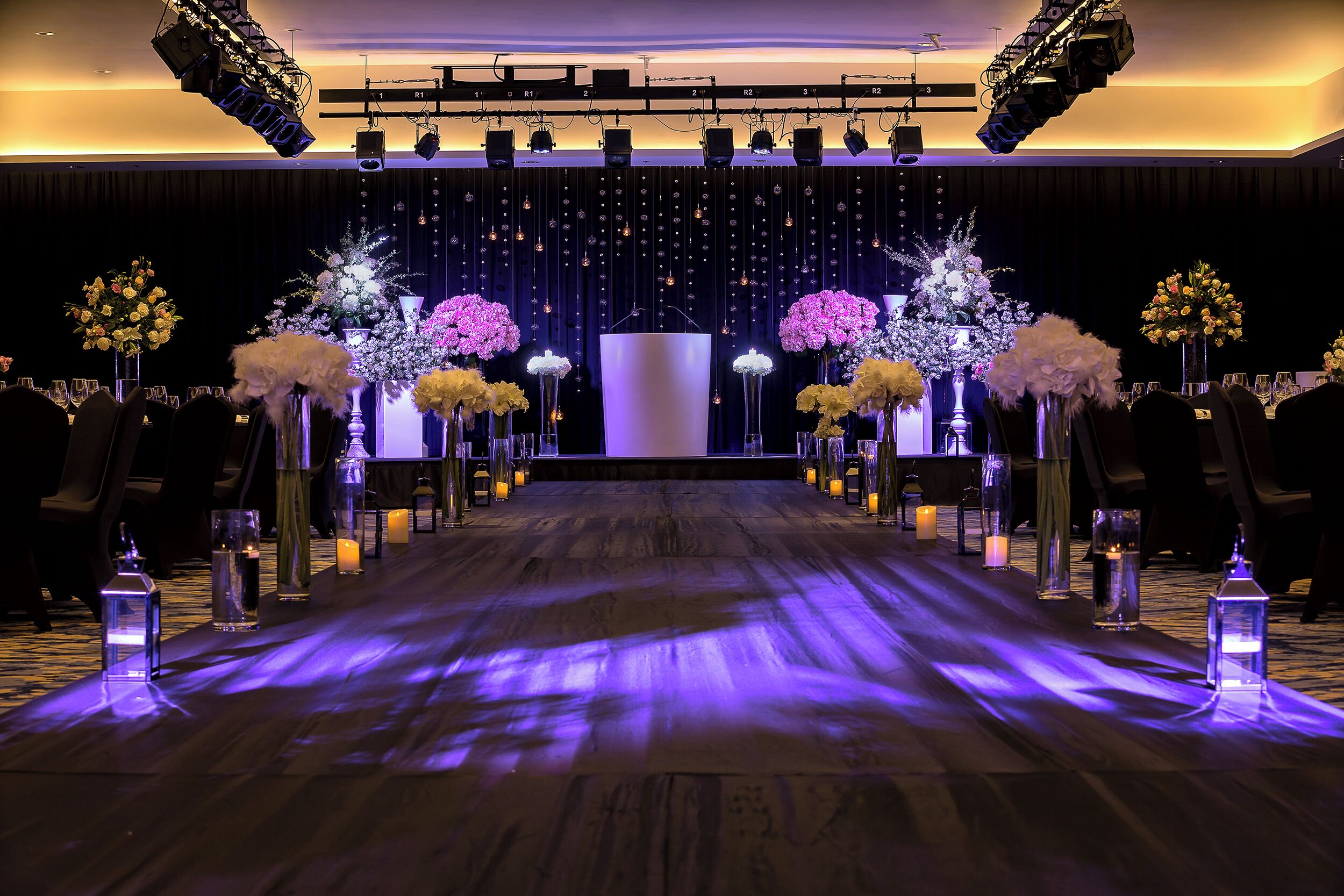 A stage with a backdrop of hanging lights and floral arrangements, a wooden floor with a purple light strip, and rows of chairs on either side.