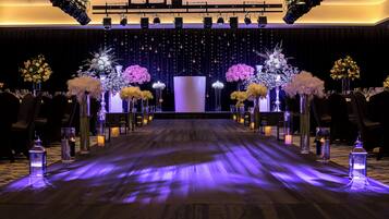 A stage with a backdrop of hanging lights and floral arrangements, a wooden floor with a purple light strip, and rows of chairs on either side.