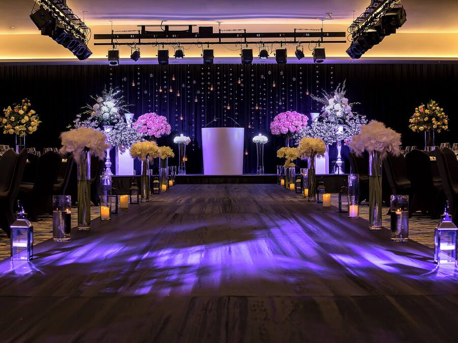 A stage with a backdrop of hanging lights and floral arrangements, a wooden floor with a purple light strip, and rows of chairs on either side.