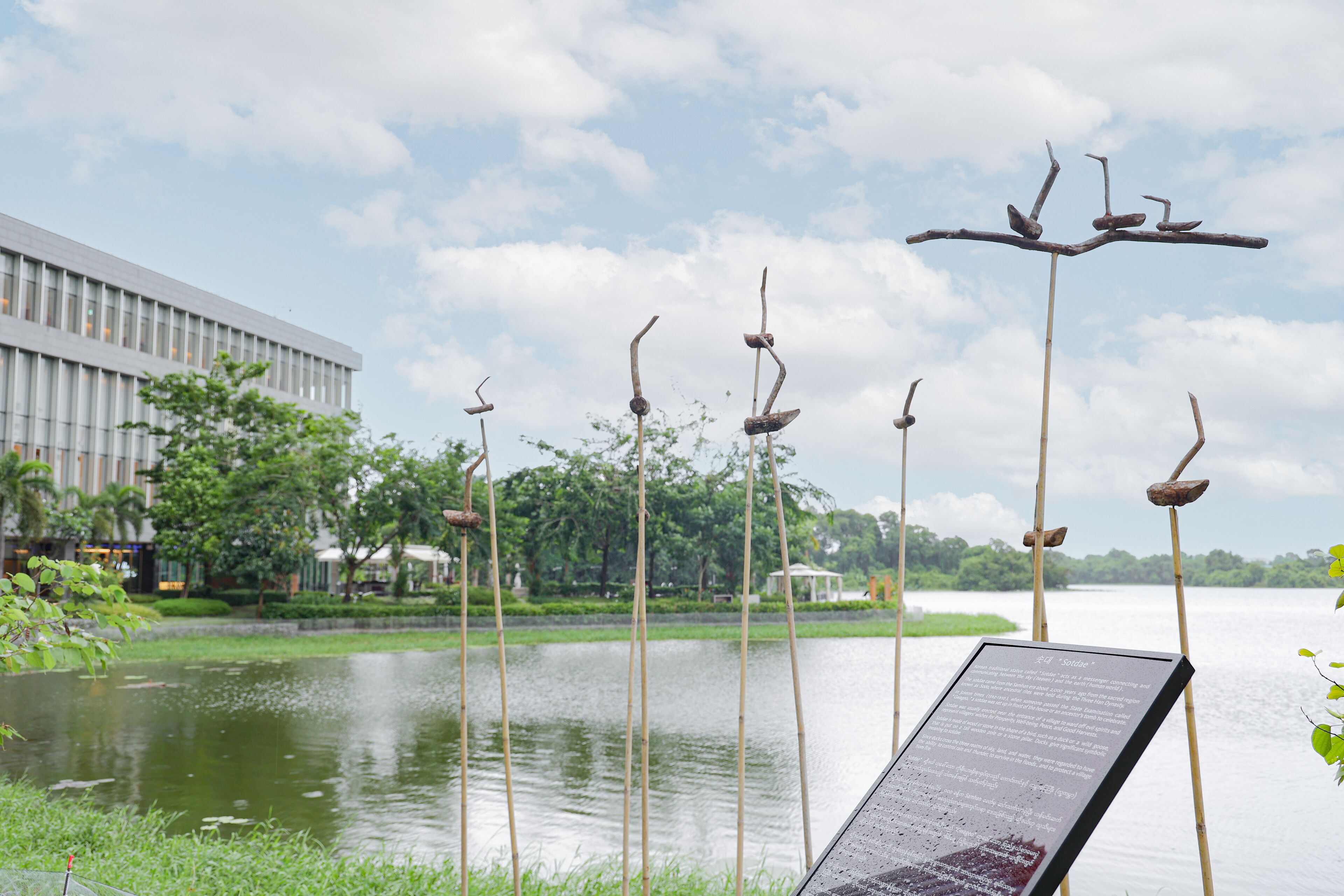 A serene lakeside view with a modern building in the background, a bamboo sculpture, and a signboard.