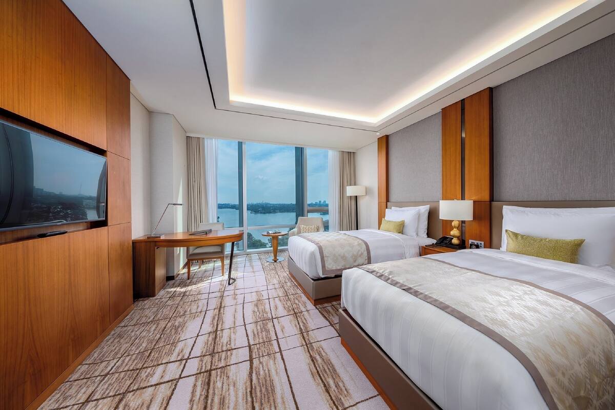 A hotel room with two beds, a TV, a desk, and a view of the city.