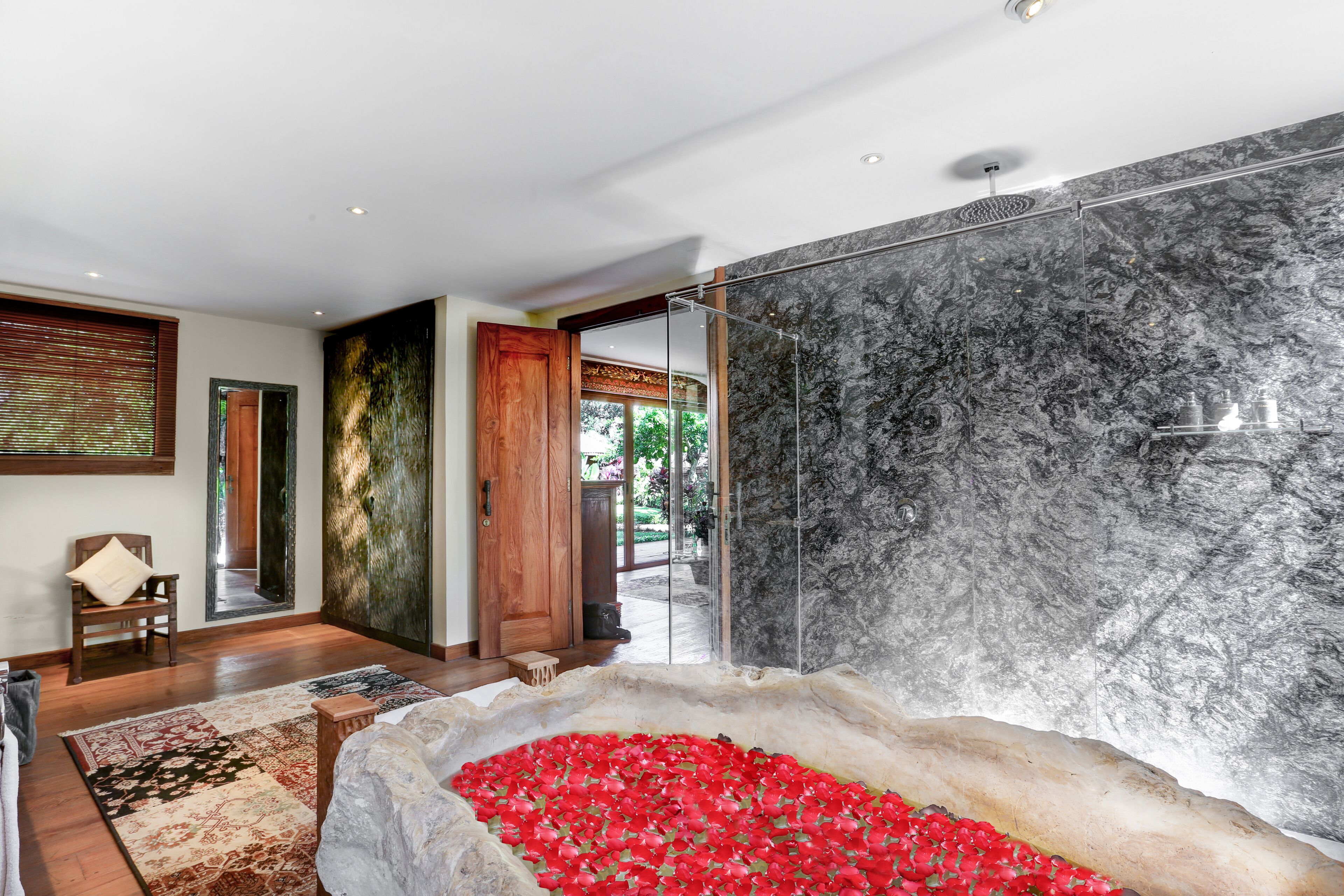 A modern bathroom with a large marble wall, a bathtub filled with red flowers, and a wooden chair.