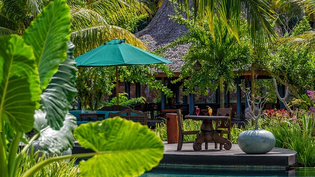 A poolside area with a table and chairs, surrounded by lush greenery and a thatched-roof structure.