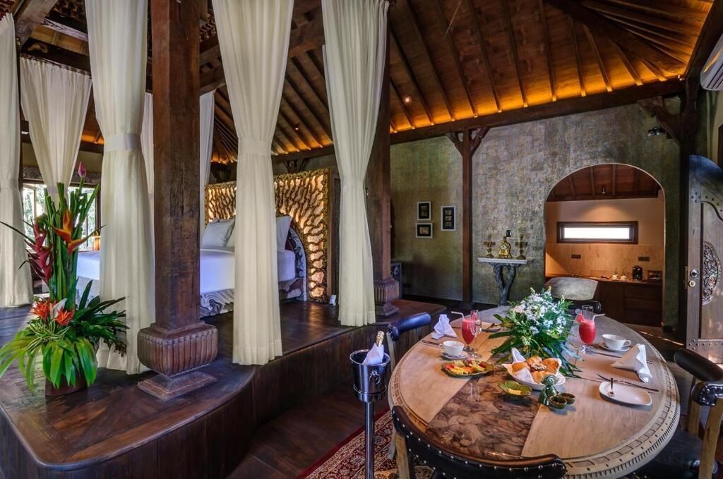 A dining area with a round table set for a meal, surrounded by wooden pillars and draped curtains.