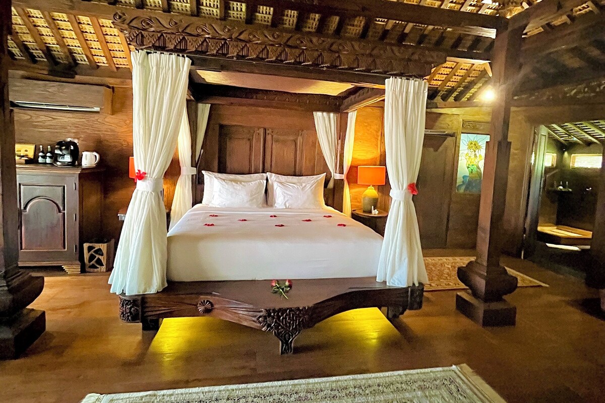 A four-poster bed with white linens and red roses, a wooden ceiling with intricate carvings, and a large ornate rug on the floor.