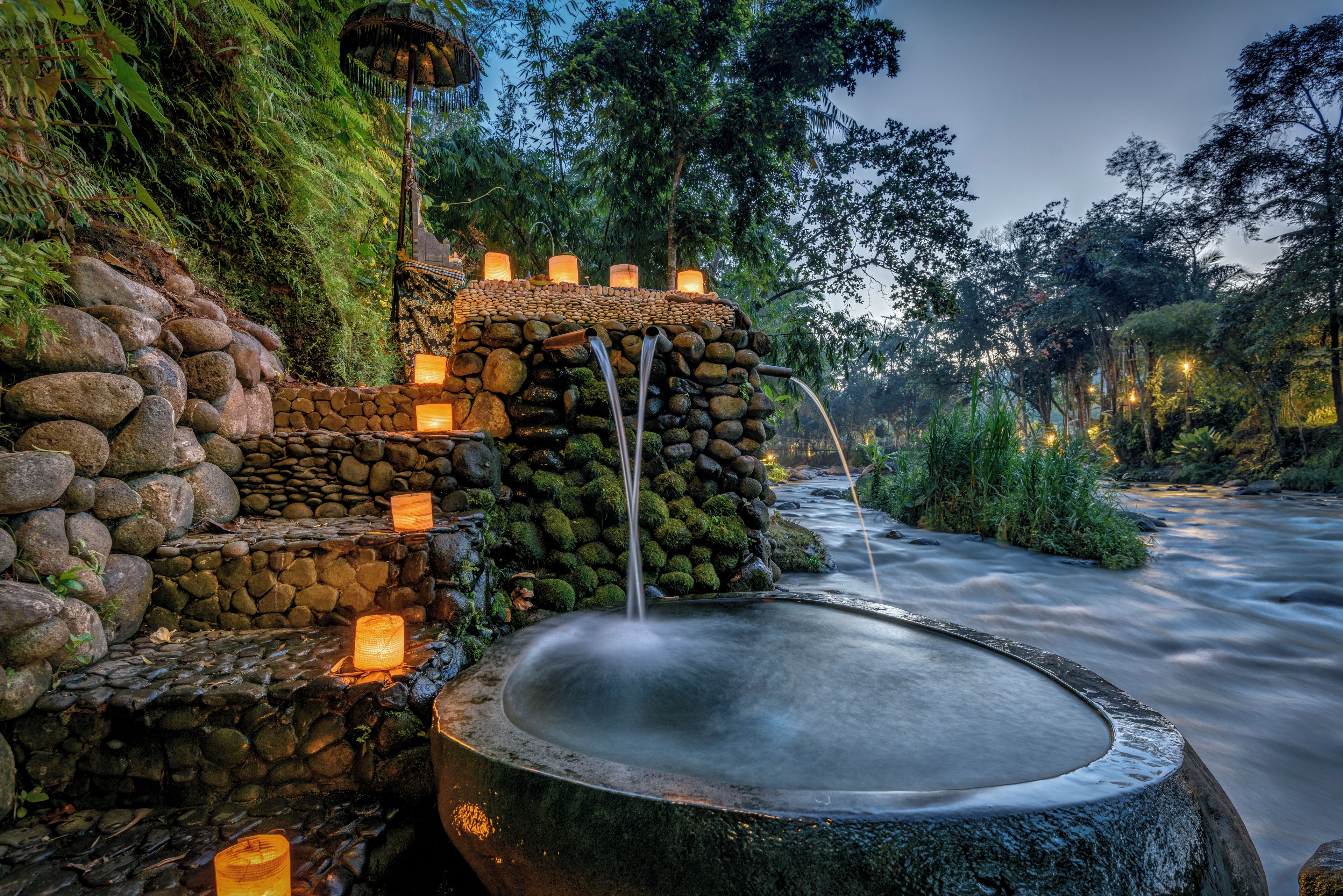 A stone water feature with cascading water, surrounded by moss-covered rocks and illuminated by lanterns.