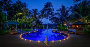 Un área de piscina tranquila con una estatua central, rodeada de bancos y velas, frente a una exuberante vegetación tropical y palmeras.