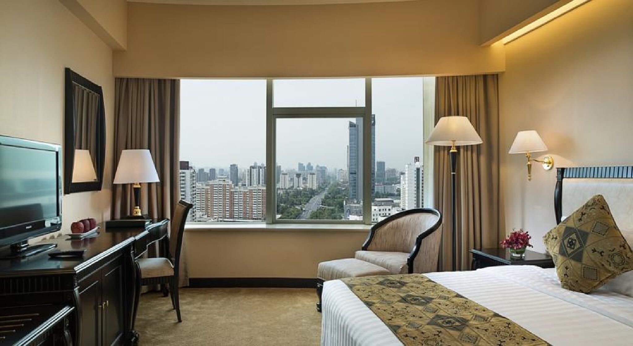 A hotel room with a bed, a desk, a chair, a TV, and a view of the cityscape.