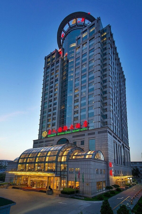 A tall hotel with a glass facade, a prominent entrance with a curved glass canopy, and a red sign indicating it is a Grand Hotel. - Celebrity International Grand Hotel (Beijing)