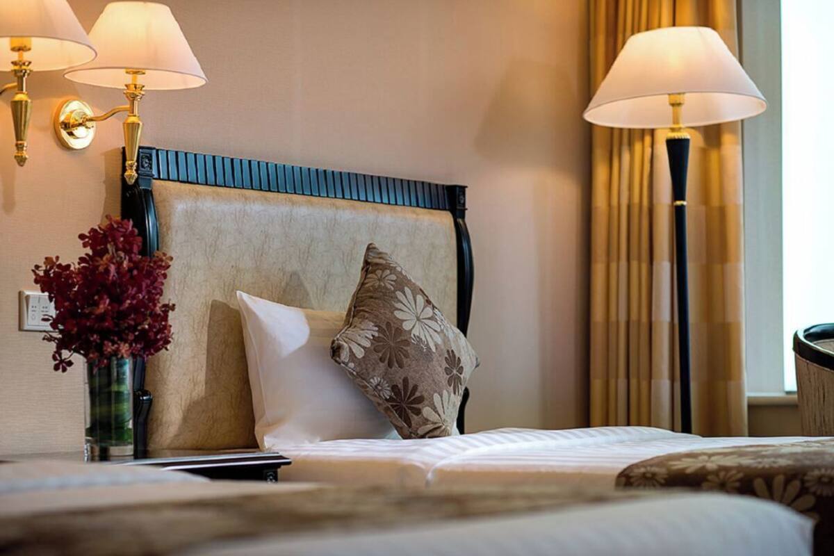 Habitación de hotel con cama, dos lámparas de pared, un jarrón con flores y una ventana con cortinas.