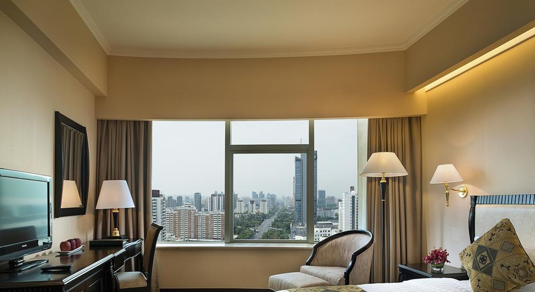 A hotel room with a large window, a desk with a television, two lamps, a chair, and a bed.