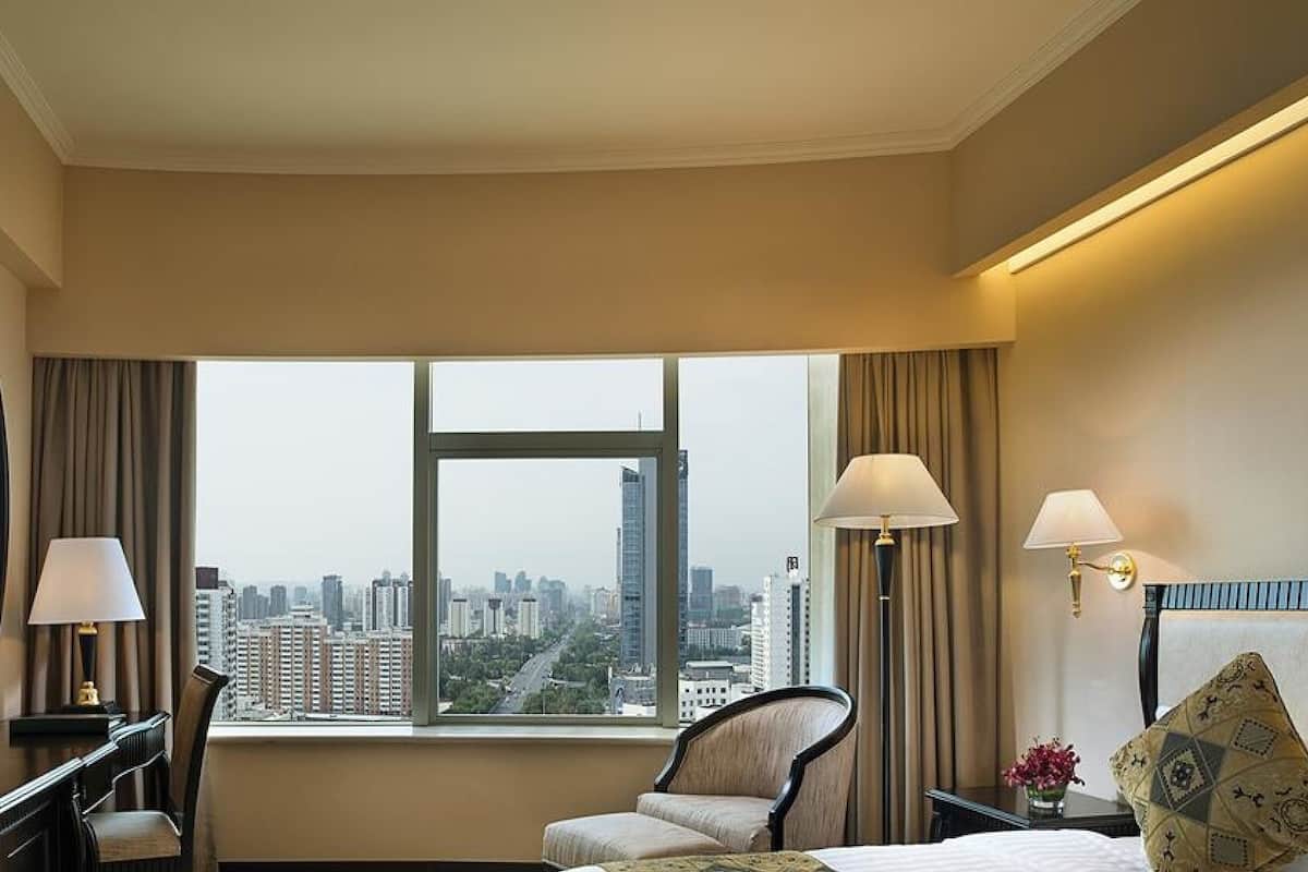 A hotel room with a large window, a desk with a television, two lamps, a chair, and a bed.