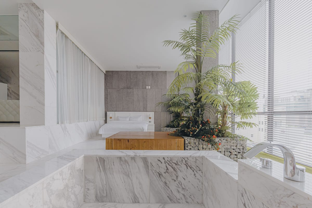 Una habitación de hotel moderna con piso de mármol, una plataforma de madera, una cama, un ventanal con persianas y una planta en maceta.