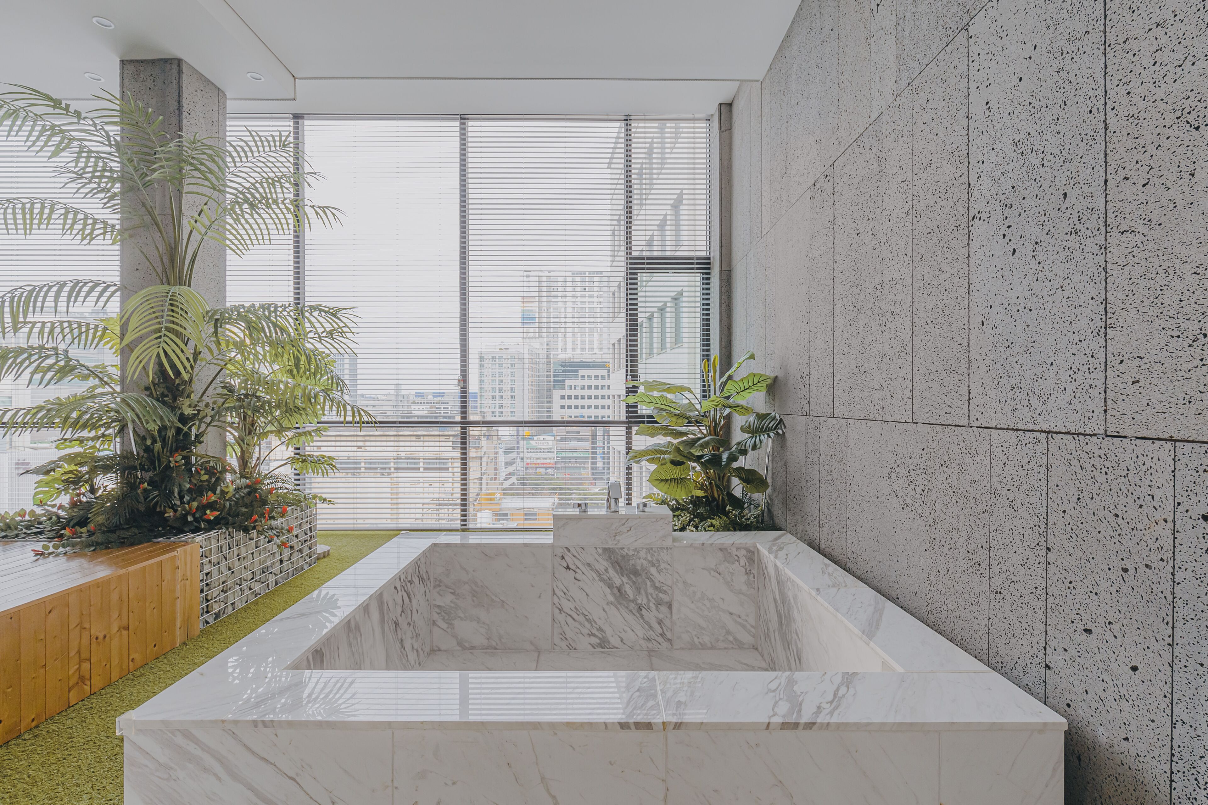 A modern bathroom with a large marble bathtub, potted plants, and a city view through large windows.