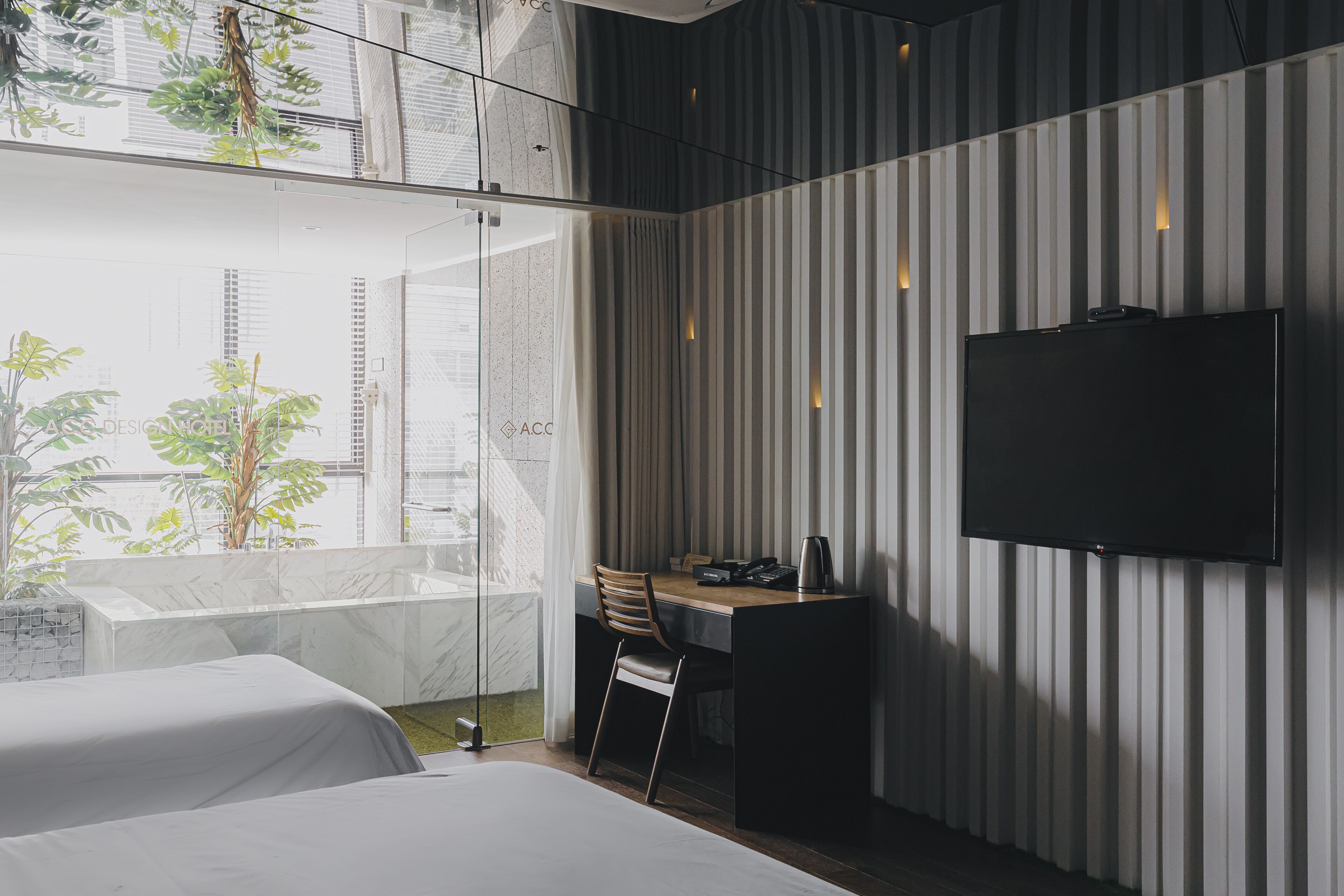 A modern hotel room with a large bed, a desk with a chair, a flat-screen TV mounted on the wall, and a view of the city through large windows.
