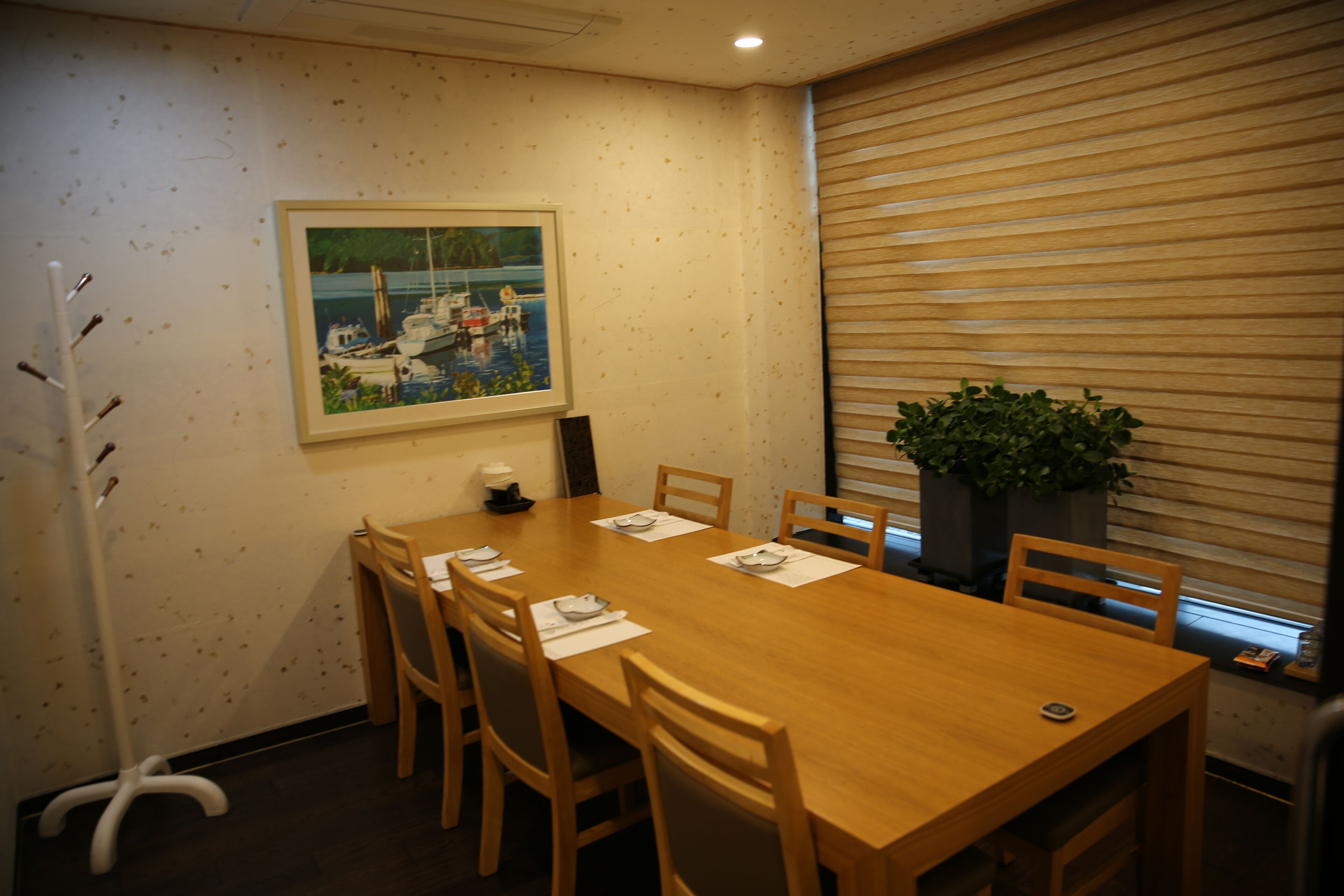 Un salón de conferencias con una mesa de madera, seis sillas, un cuadro de barcos en la pared, una planta en maceta y una ventana con persianas.