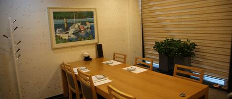 Un salón de conferencias con una mesa de madera, seis sillas, un cuadro de barcos en la pared, una planta en maceta y una ventana con persianas.