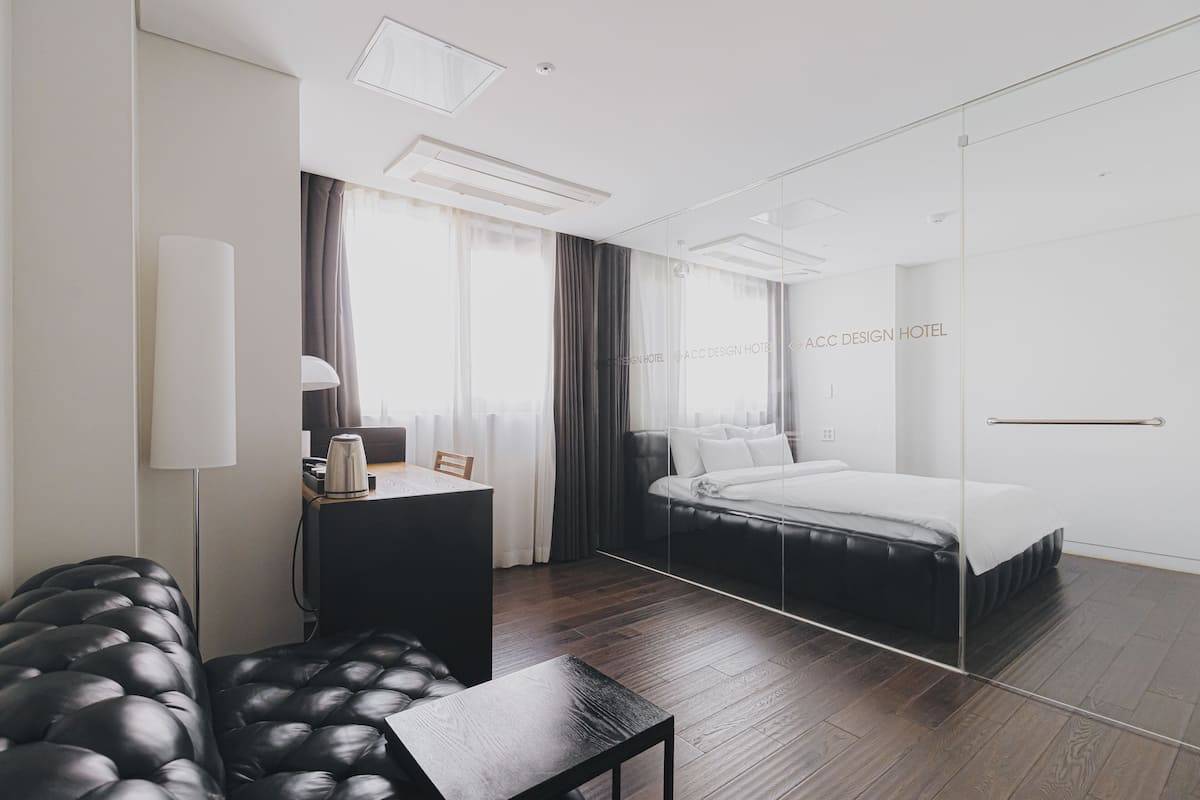 Una habitación de hotel moderna con una cama grande, un escritorio con lámpara, un sofá de cuero y un muro divisorio de vidrio.