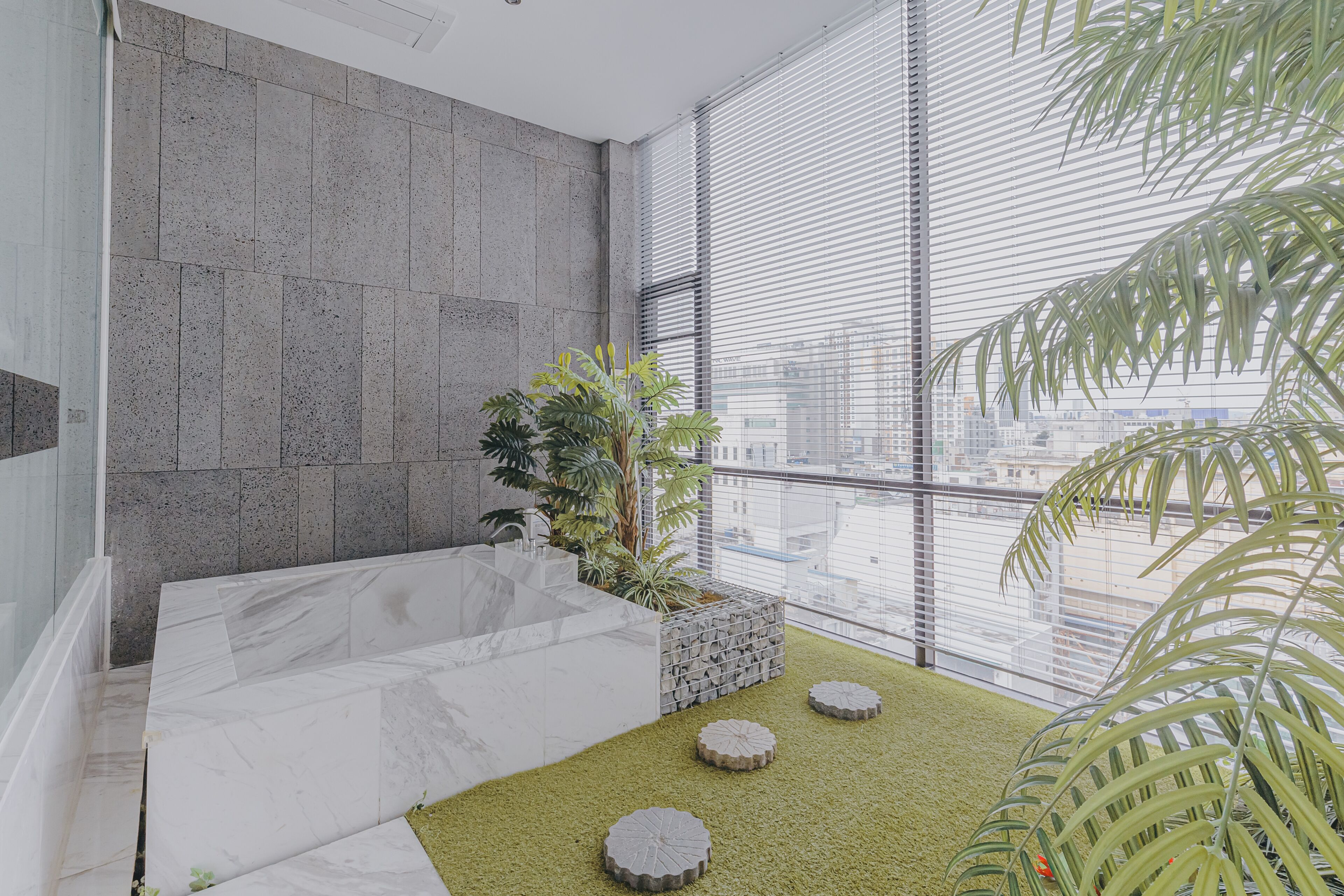 A modern bathroom with a marble bathtub, a small plant, and a view of the city through large windows.