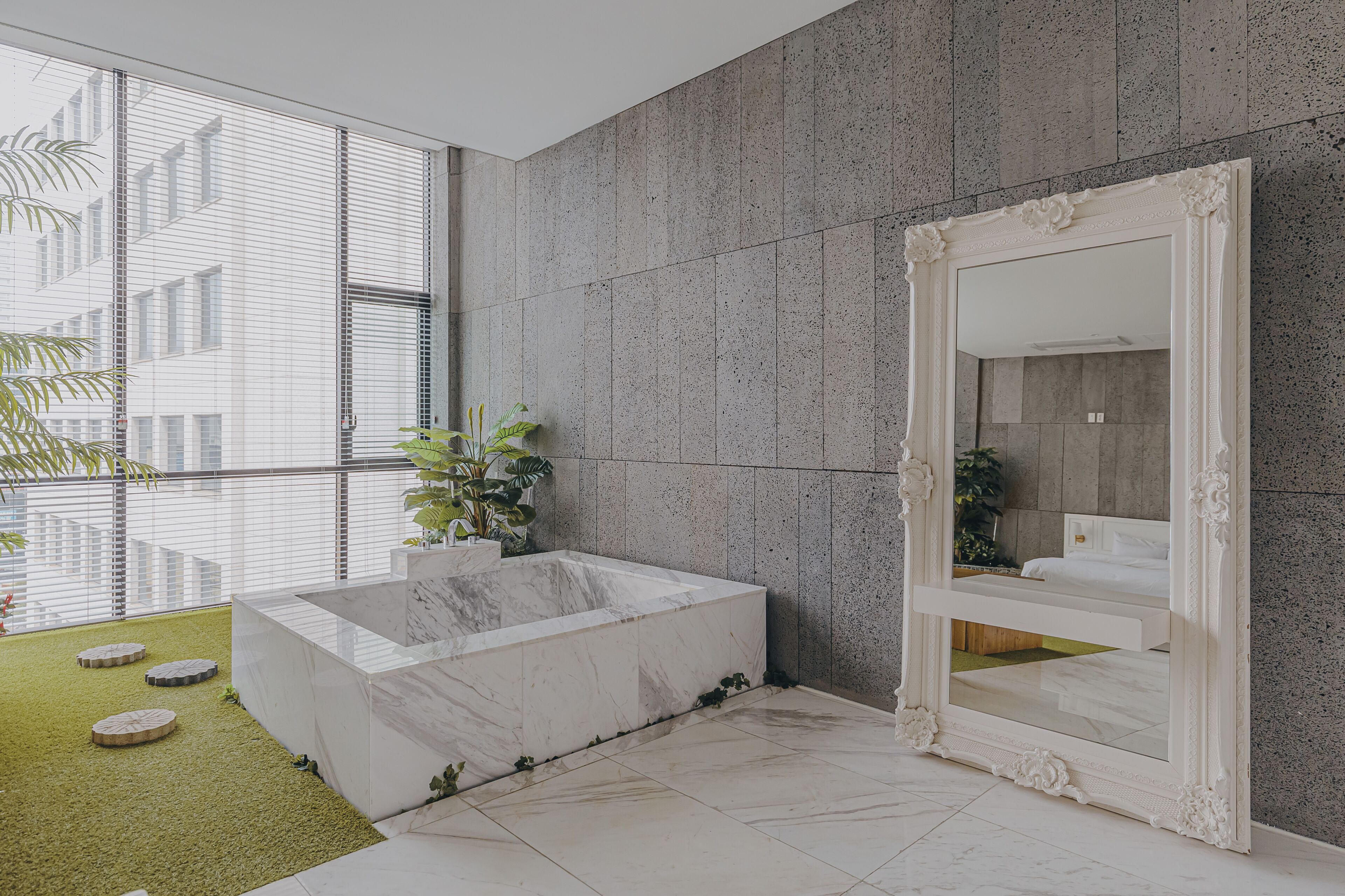 A modern bathroom with a marble bathtub, a large mirror, and a view of the city through large windows.