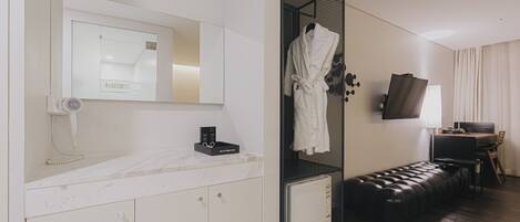 Habitación de hotel con un espejo grande, un tocador con encimera de mármol, una bata colgada en un perchero, un mueble pequeño, un banco de cuero negro, un escritorio con lámpara y un televisor en la pared.